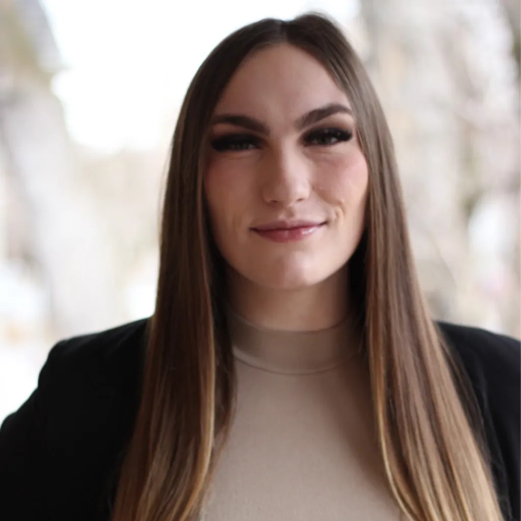 Headshot of Lived Experience (LEx) Leader Natalie Clark smiling, wearing a black blazer over a beige turtleneck, with long straight brown hair with highlights, against a blurred outdoor background.
