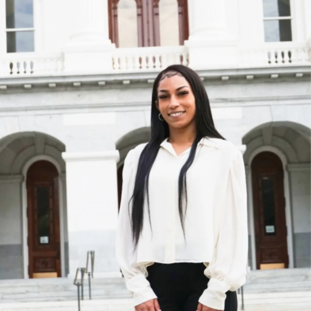 Headshot of Lived Experience (LEx) Leader Jasmine Green smiling, wearing a white blouse and black pants, with long straight black hair, standing in front of a large white building.