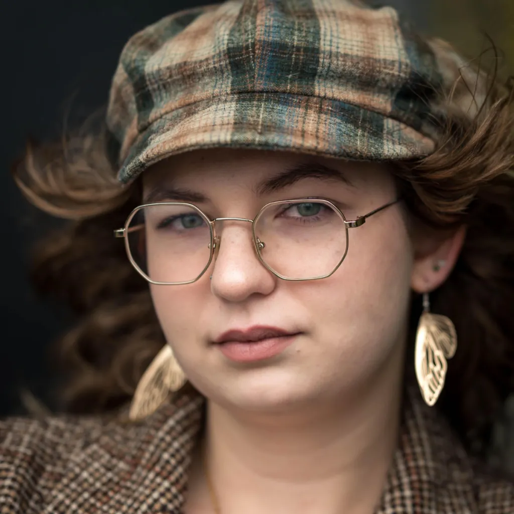 Headshot of Lived Experience (LEx) Leader Charissa Keebaugh with auburn hair blowing in the wind, wearing a plaid newsboy cap, gold octagonal glasses, gold leaf earrings, and a tweed jacket, against a dark background.