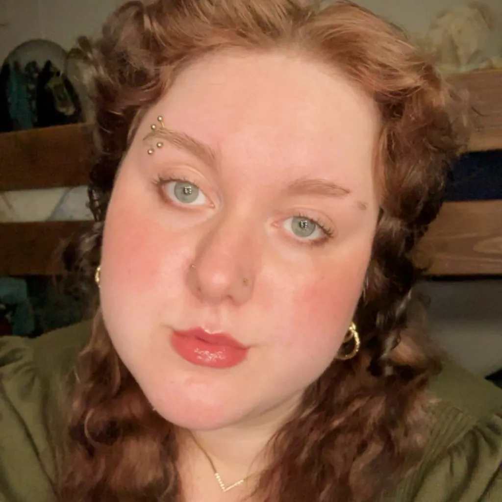 Headshot of Brina Williams wearing an olive green top, gold hoop earrings, and a delicate necklace, with curly auburn hair, multiple eyebrow piercings, a nose ring, and red lipstick, in an indoor setting.