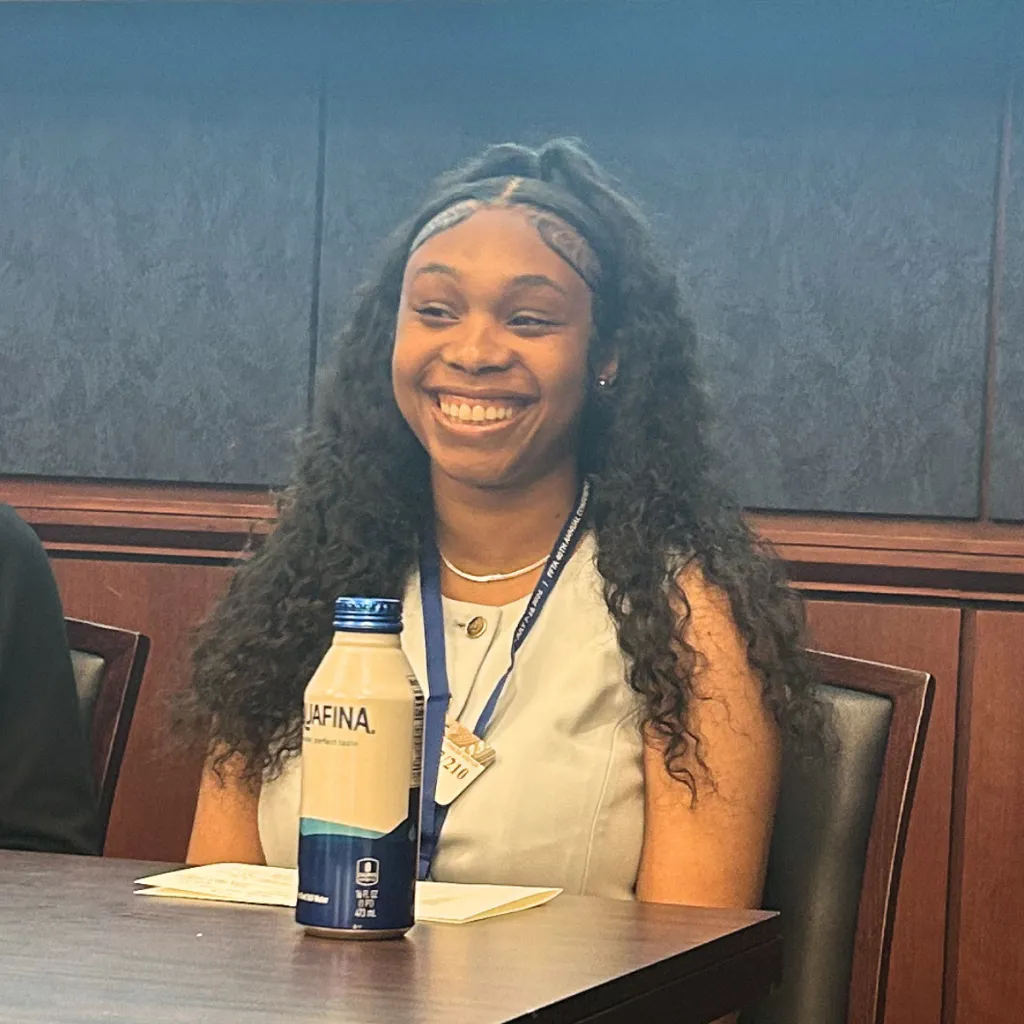 FosterClub LEx Leader Knowledge Grant smiling broadly while seated at a conference table, wearing a conference lanyard and a cream top, in a formal meeting room setting.